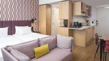 A modern hotel room with a large bed, a sofa with a yellow pillow, a kitchenette, and a dining area.