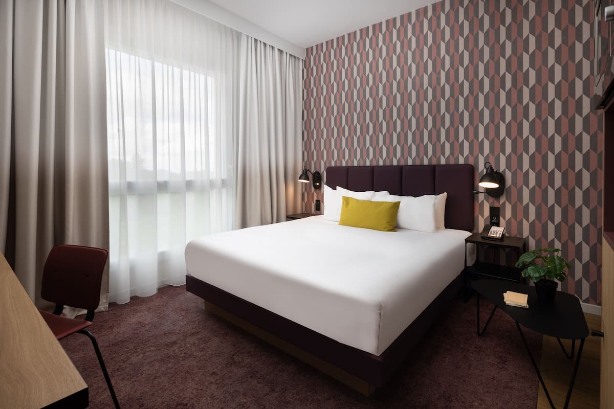 Une chambre d’hôtel avec un grand lit, un bureau, une chaise, une table de chevet avec une lampe et une plante en pot.