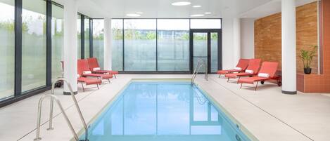 A modern indoor swimming pool with clear glass walls, red lounge chairs, and a wooden accent wall.