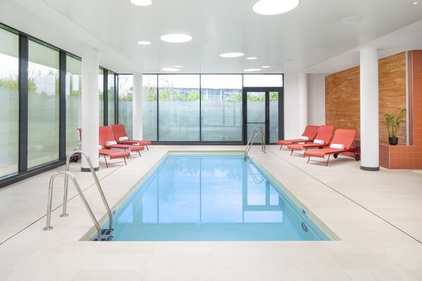 A modern indoor swimming pool with clear glass walls, red lounge chairs, and a wooden accent wall.