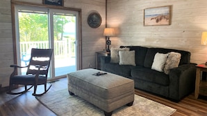 Ein gemütliches Wohnzimmer mit einem Schaukelstuhl, einem grauen Hocker und einem dunkelgrauen Sofa mit gemusterten Kissen. Ein spiegel mit Holzrahmen hängt an der Wand, und eine Lampe steht auf einem Beistelltisch.