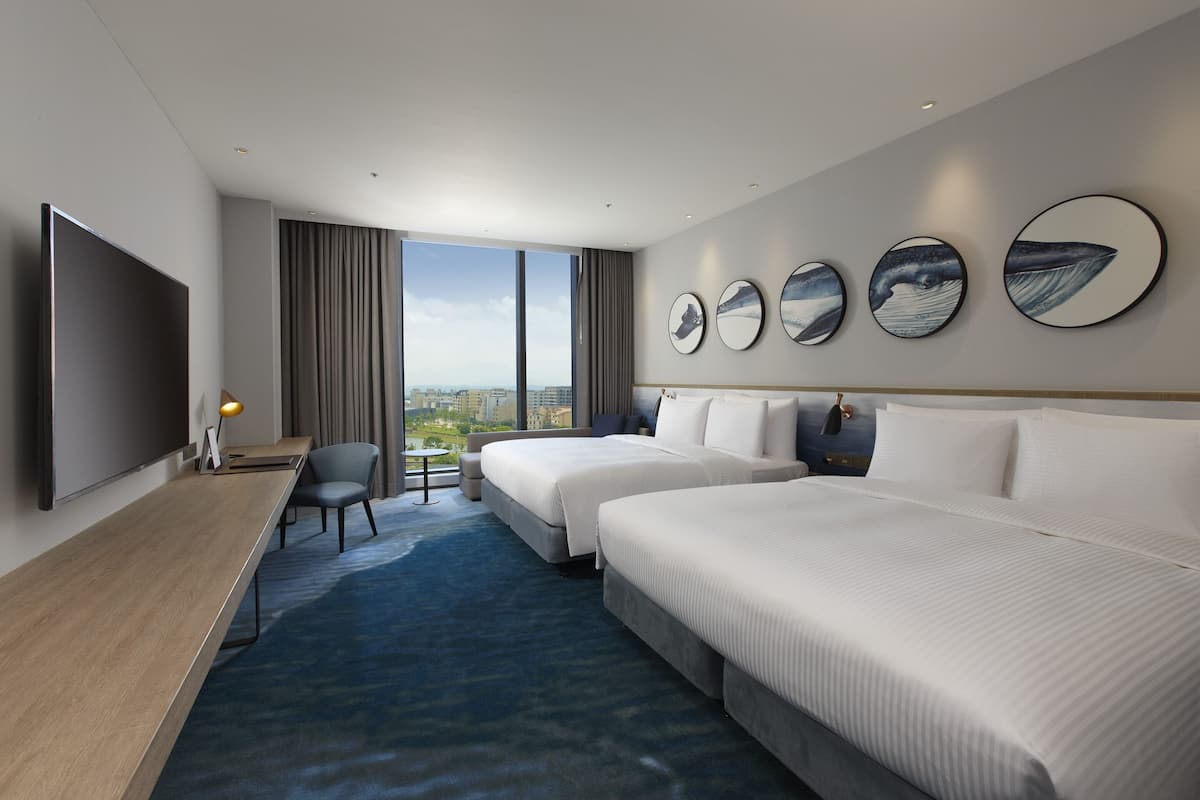 Habitación de hotel con dos camas, un escritorio, televisor y una ventana grande con vistas a la ciudad.