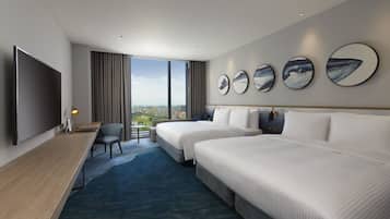 A hotel room with two beds, a desk, a TV, and a large window with a city view.