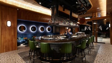 Zona de bar moderna con una barra curvada, taburetes de bar verdes y un área de estar con un sofá tapizado en azul.