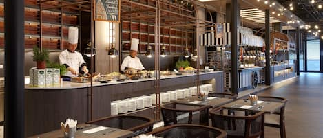 A restaurant with a modern interior, featuring a counter with chefs preparing food, neatly arranged tables with set dining ware, and a variety of dishes displayed on shelves.