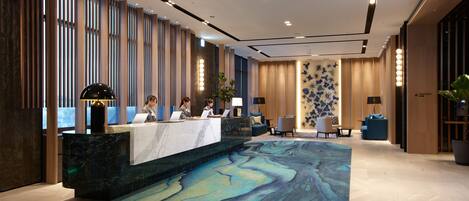 A hotel reception area with a marble counter, three staff members, a blue patterned carpet, and a large potted plant.