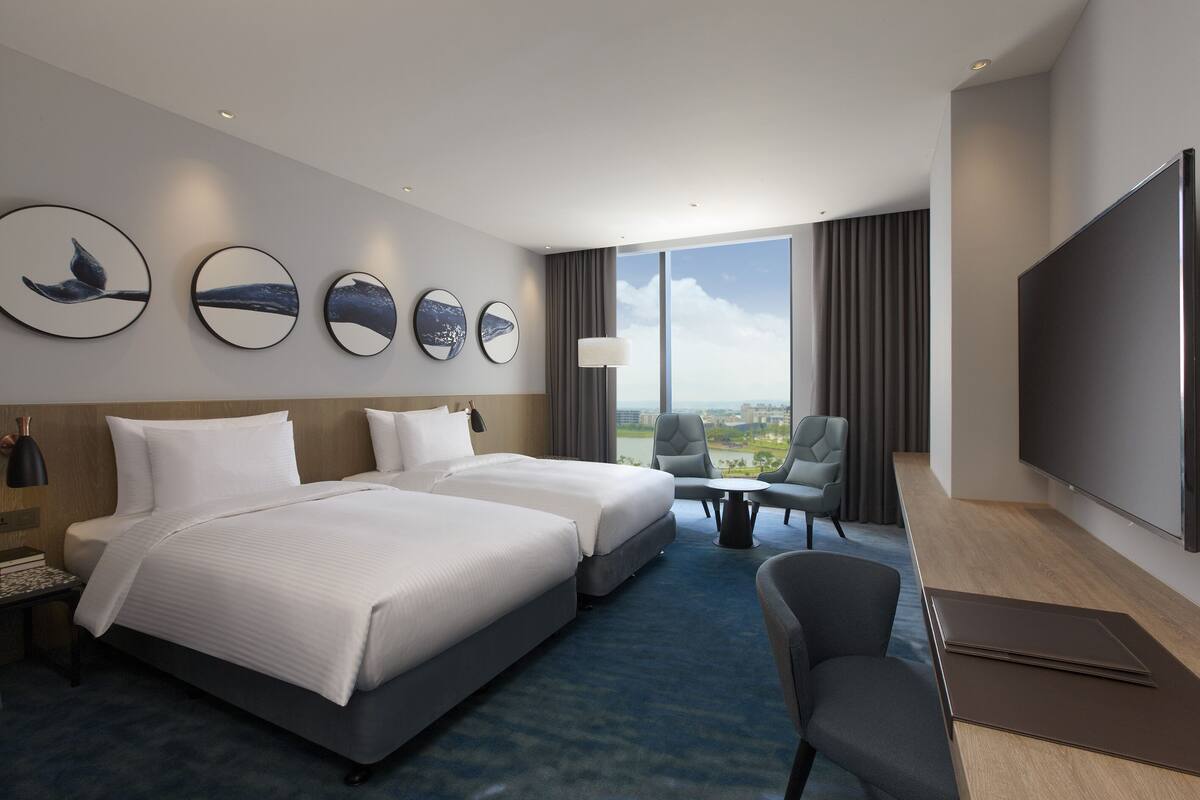 Habitación de hotel con dos camas, televisión, escritorio y vista a la ciudad.