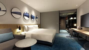 A modern hotel room with a large bed, a sofa, a desk, and a TV. There are three wall mirrors and a small round table.