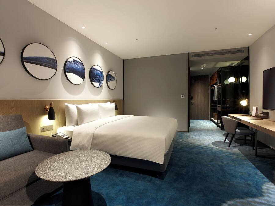 A modern hotel room with a large bed, a sofa, a desk, and a TV. There are three wall mirrors and a small round table.