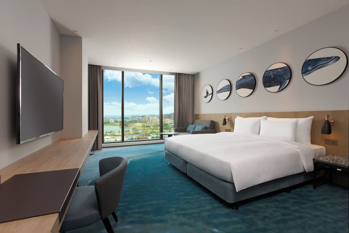 Habitación de hotel moderna con una cama grande, un escritorio, una silla y un televisor. Amplias ventanas ofrecen vistas a la ciudad.