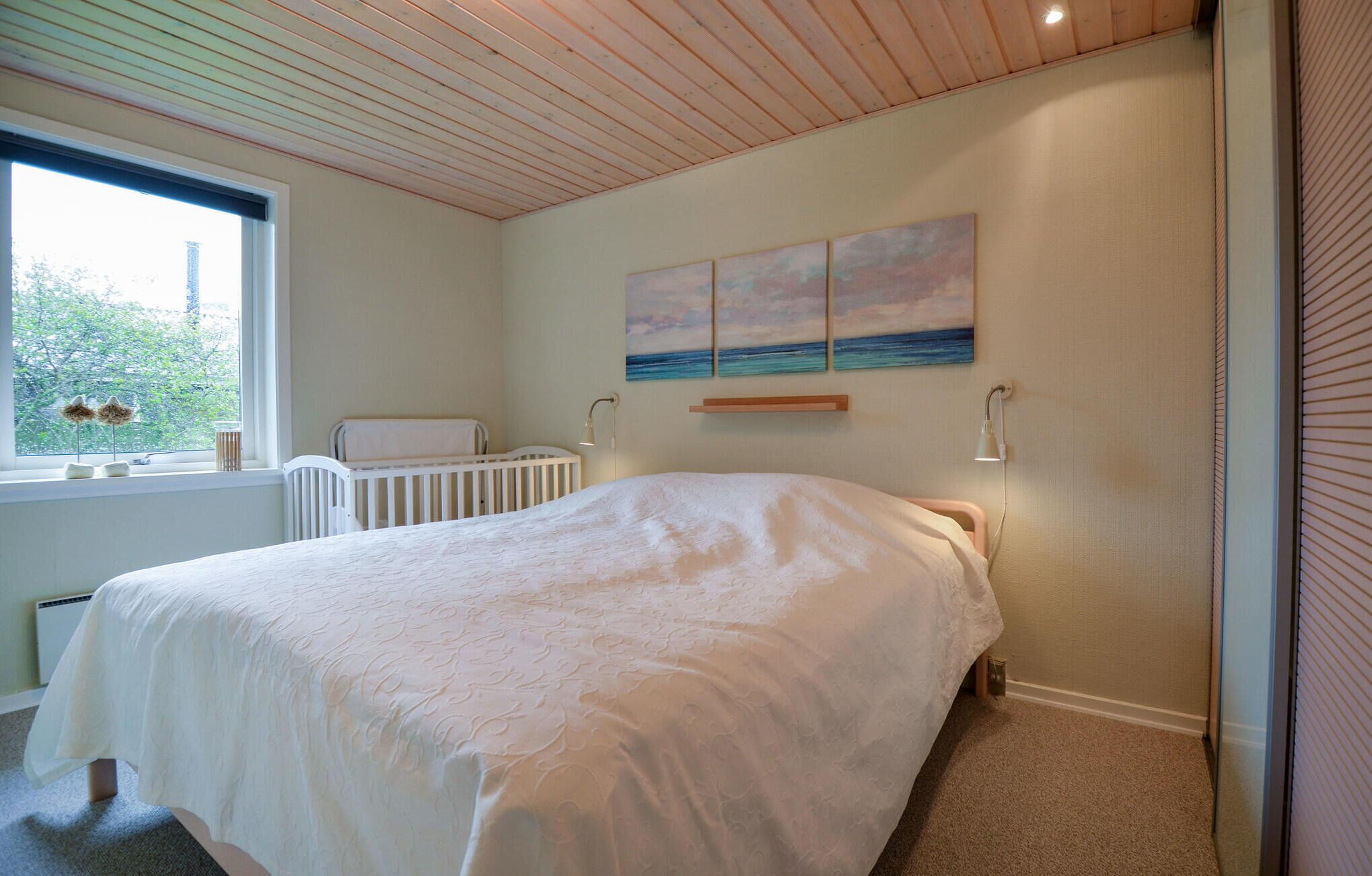 A bedroom with a large bed, a crib, a window with a view, and a painting on the wall.