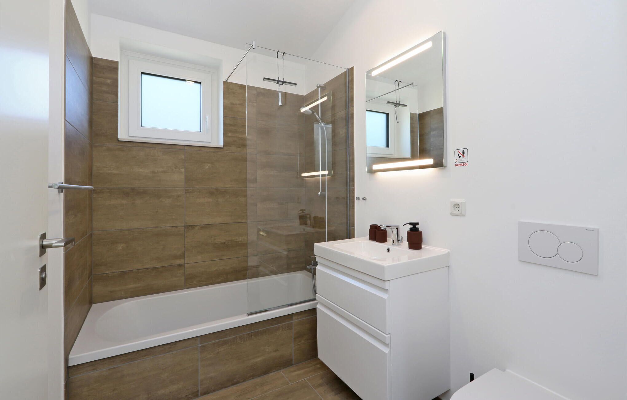 A modern bathroom with a glass-enclosed shower, a white sink with a mirror above it, and a wooden floor.