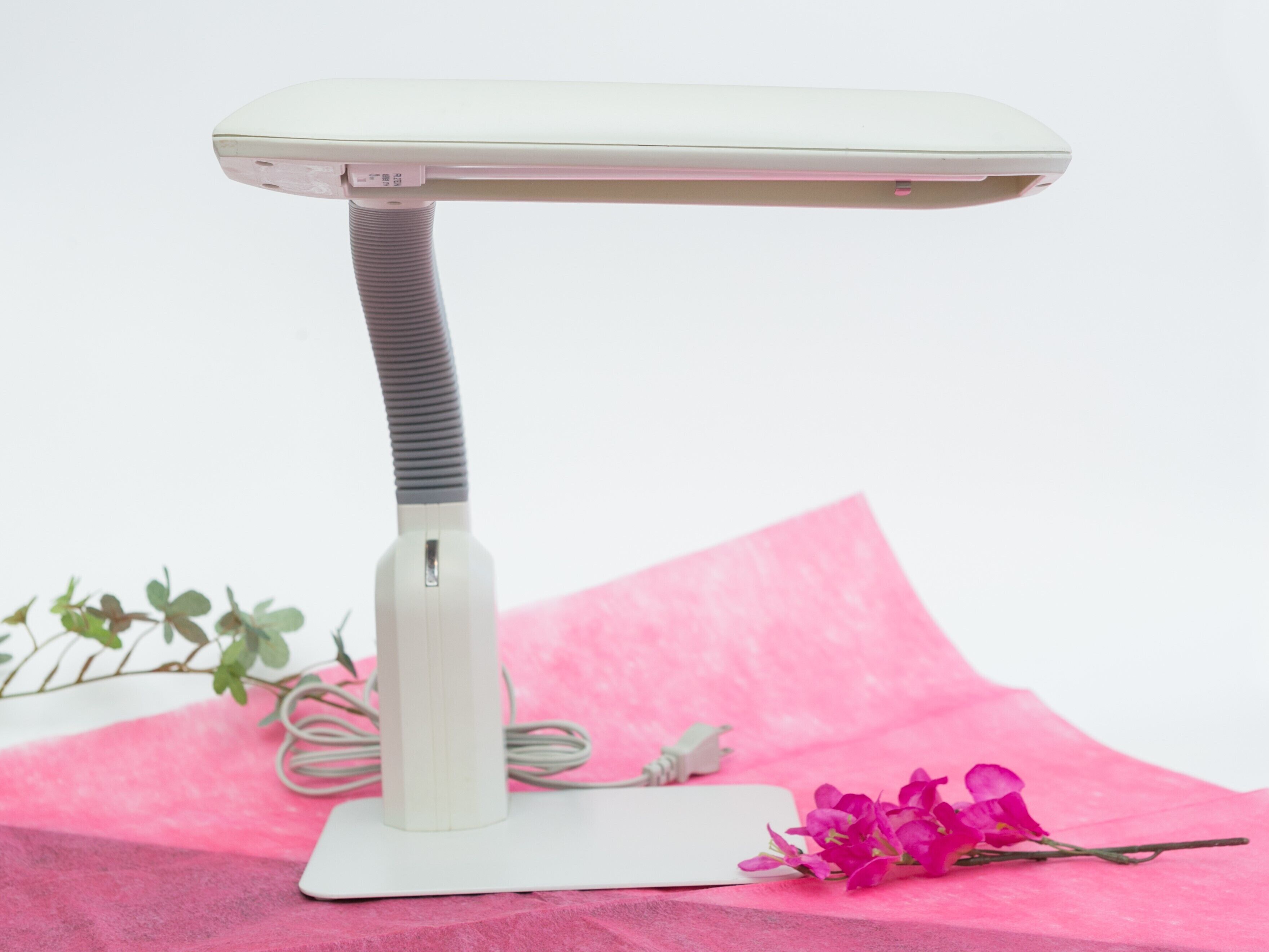 A white portable heater with a flexible neck, placed on a pink surface with a flower and a plant in the background.