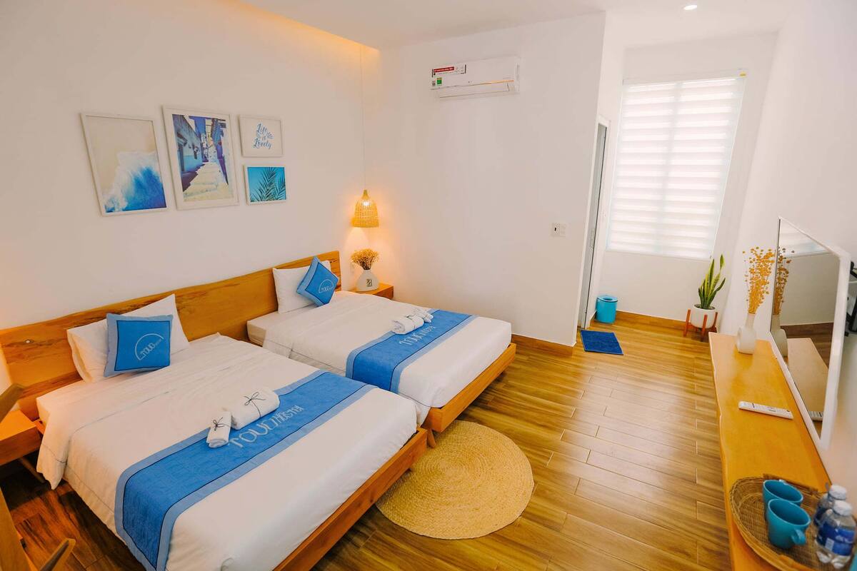 A hotel room with two beds, a wooden floor, a round rug, a small table with a water bottle, and a wall-mounted air conditioner.
