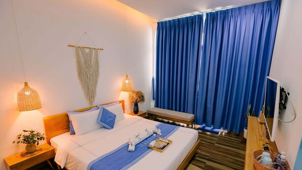 A neatly arranged hotel room with a bed, bedside lamps, a bench, a desk, and a large window with curtains. - To Vo Hue Hostel (Hue)