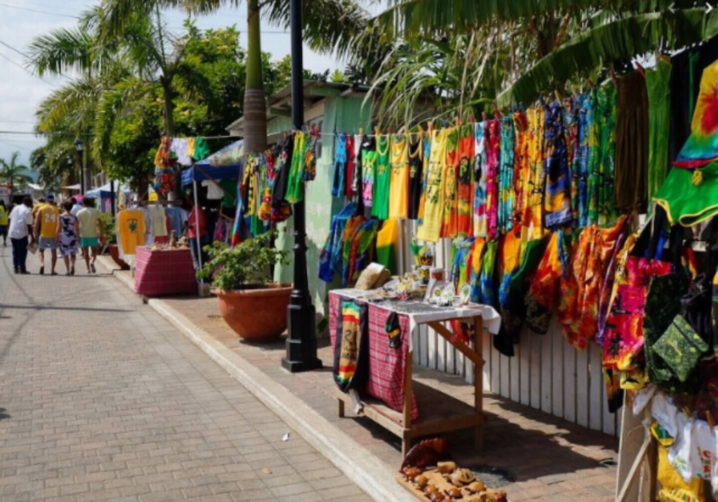 Un marché de rue où l’on peut admirer des étoffes aux couleurs vives, disposées en tentures pour la vente, une petite table où sont exposés divers objets, et des palmiers en arrière-plan.