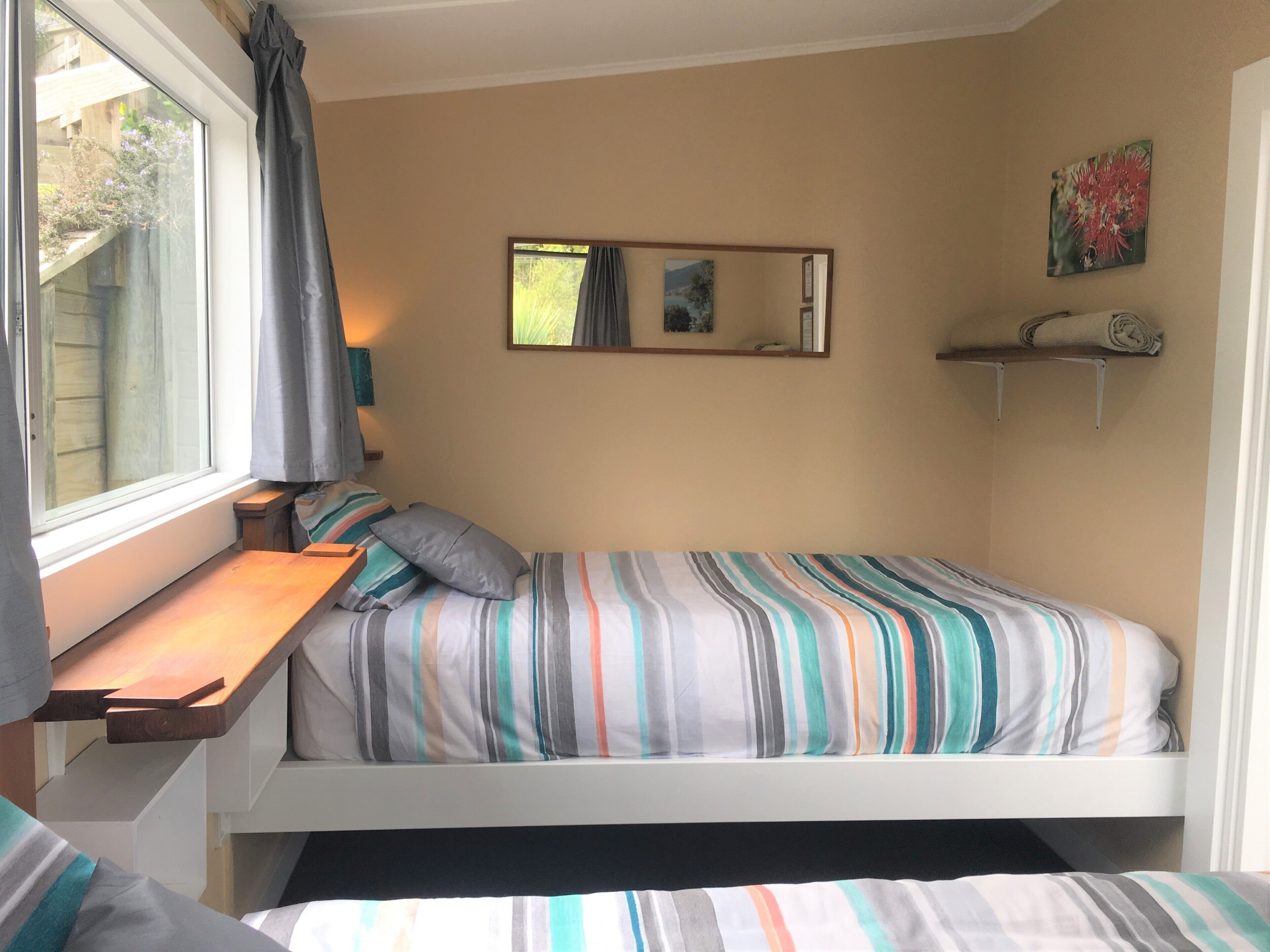 A bedroom with a bed, a window with curtains, a bedside table, a mirror, and a wall-mounted shelf.