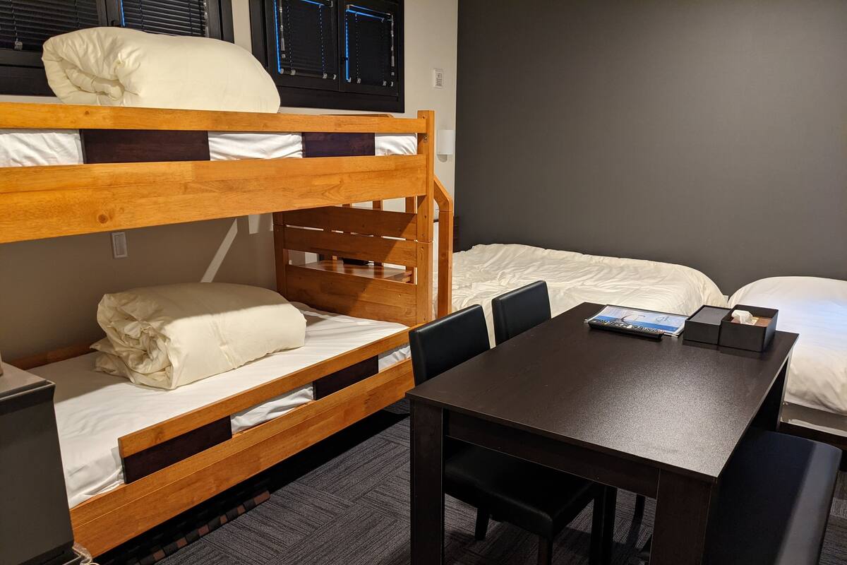 Une chambre avec un lit superposé, un bureau et des chaises, une petite table et une fenêtre avec des stores.