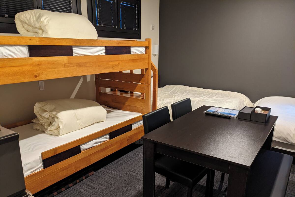 Une chambre avec un lit superposé, un bureau et des chaises, une petite table et une fenêtre avec des stores.
