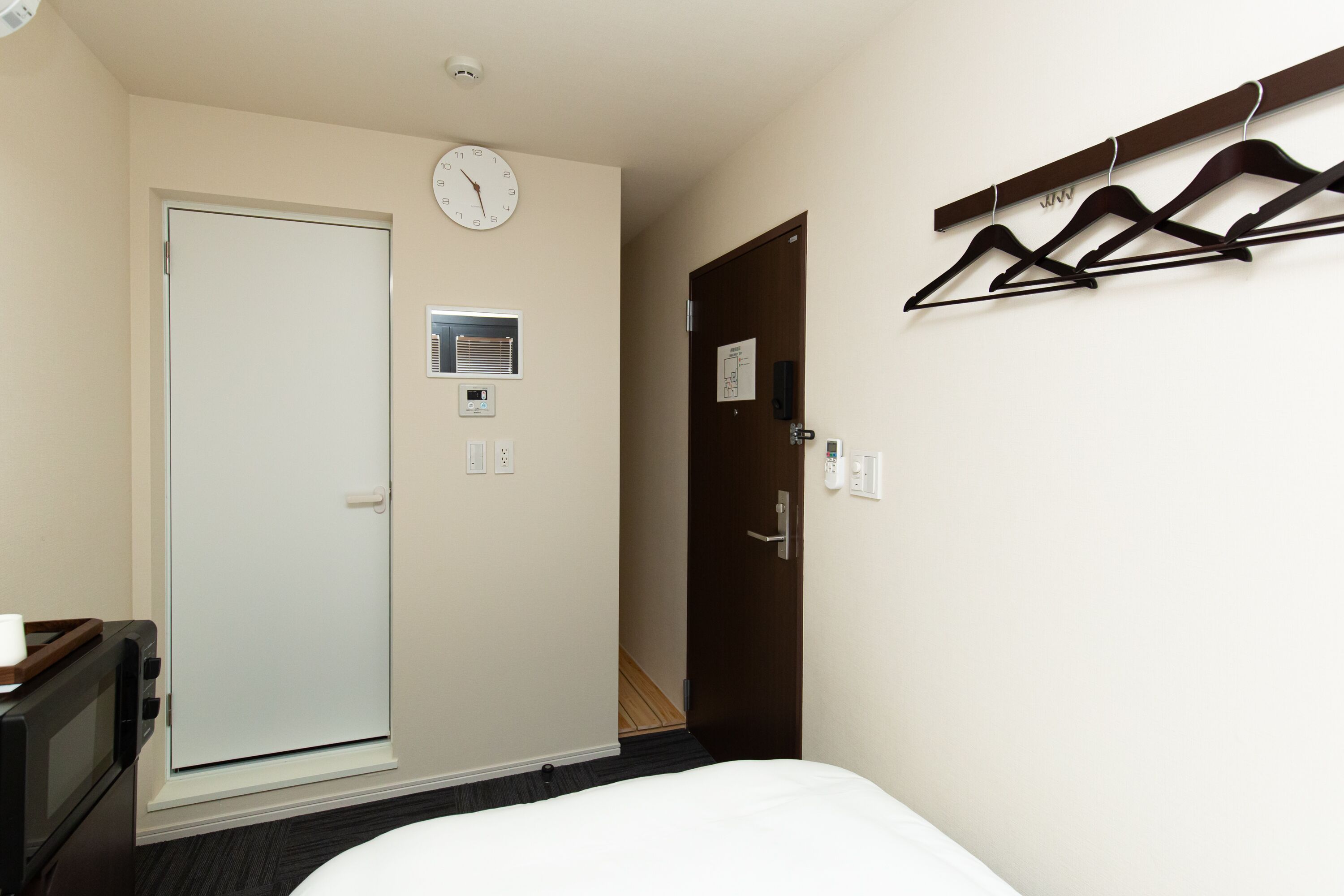 A hotel room with a bed, a white door, a clock, a coat rack, and a small window.