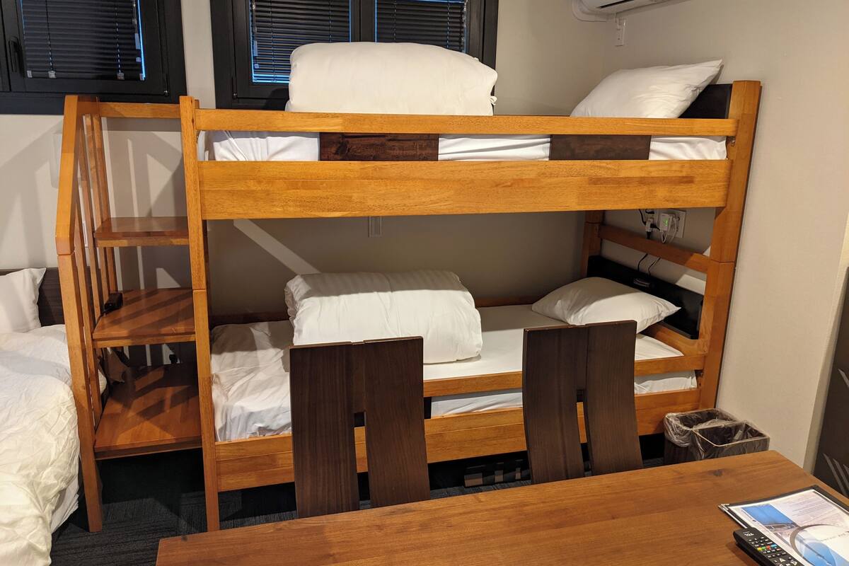 Un lit superposé avec une structure en bois, du linge de lit blanc et une table avec une télécommande.