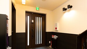A hotel lobby with a dark wooden door, a reception desk, a fire alarm, and a wall-mounted light fixture. - Gennoya (Tokyo)