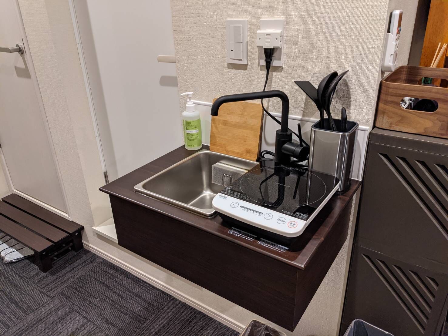 A compact kitchenette with a built-in sink, a small countertop, a bottle of hand soap, and a power outlet.