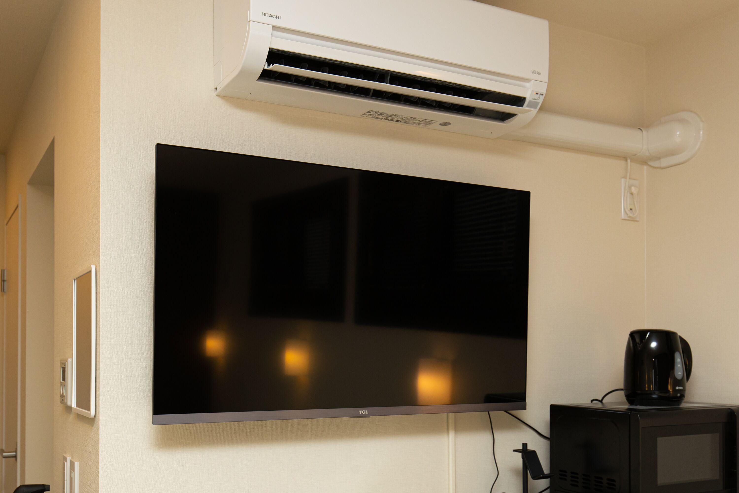 A flat-screen TV mounted on a wall, an air conditioning unit above it, and a small black appliance on a cabinet below.