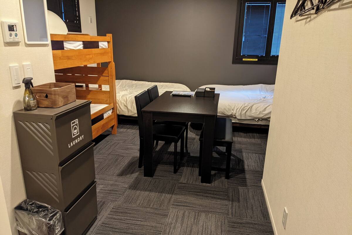 Une chambre équipée d’un lit superposé, d’un bureau en bois, d’une chaise, d’une petite poubelle et d’un espace dédié au linge sale.