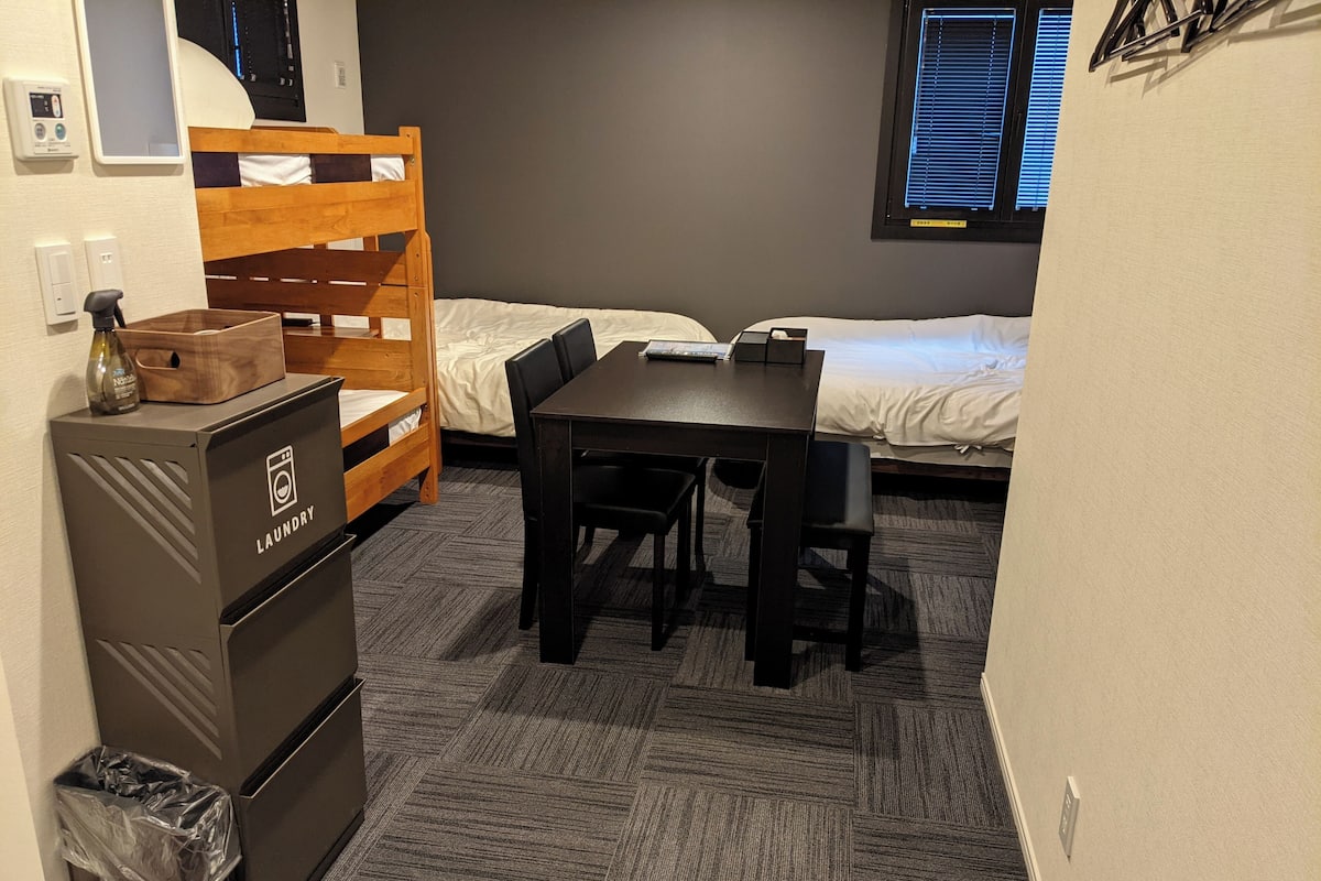 A room with a bunk bed, a wooden desk, a chair, a small trash bin, and a laundry station.