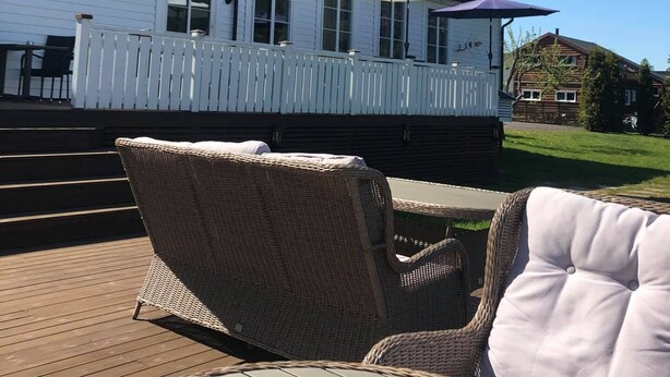 A wooden deck with wicker chairs and a white cushioned chair. A white fence and a building with a black roof are visible in the background. - Dølen Hotel (Evje)