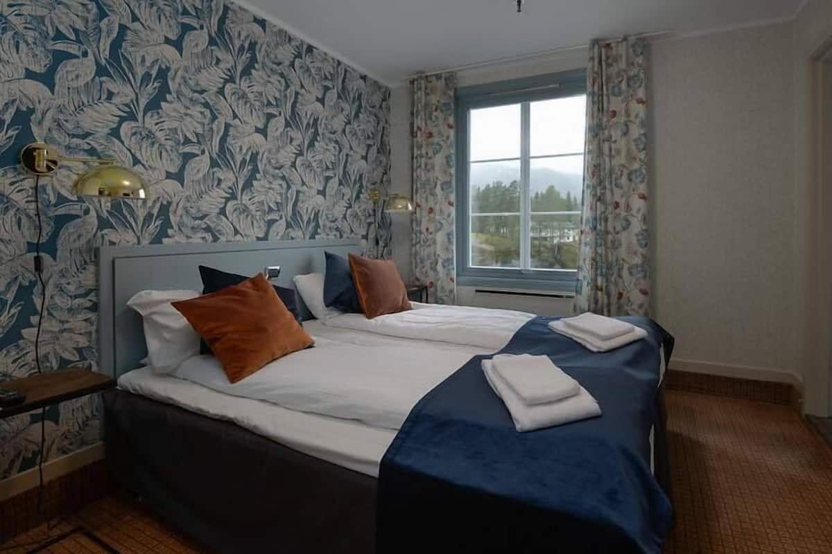 Habitación de hotel con una cama grande, dos mesitas de noche, una ventana con cortinas y una pared con estampado floral.