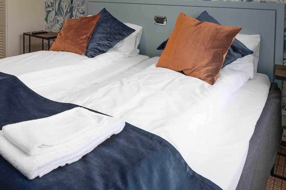 Cama doble con ropa de cama blanca y azul marino, dos almohadas y una toalla doblada.