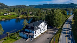A large, white building with a black roof, likely a hotel, situated beside a lake with a road running parallel to it. - Dølen Hotel (Evje)
