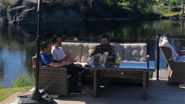 A group of people seated on a wicker sofa and chairs on a wooden deck by a lake, with a large umbrella providing shade. - Dølen Hotel (Evje)