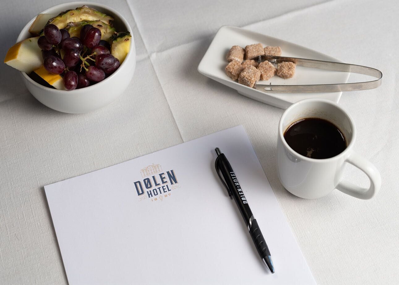 A white table with a bowl of fruit, a plate of sugar cubes, a cup of coffee, a pen, and a paper with hotel branding.