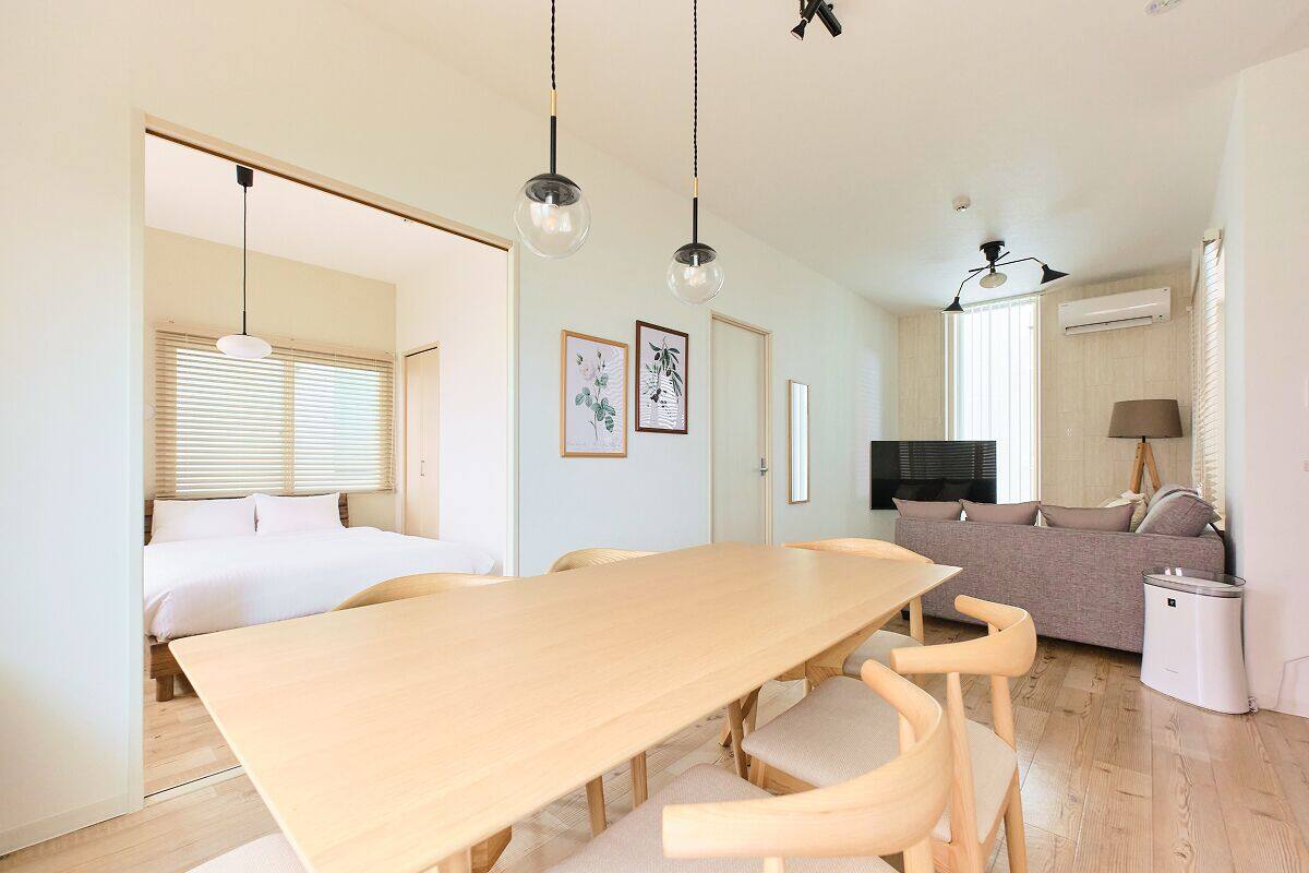 A modern, well-lit room with a wooden dining table, chairs, a sofa, and a TV.