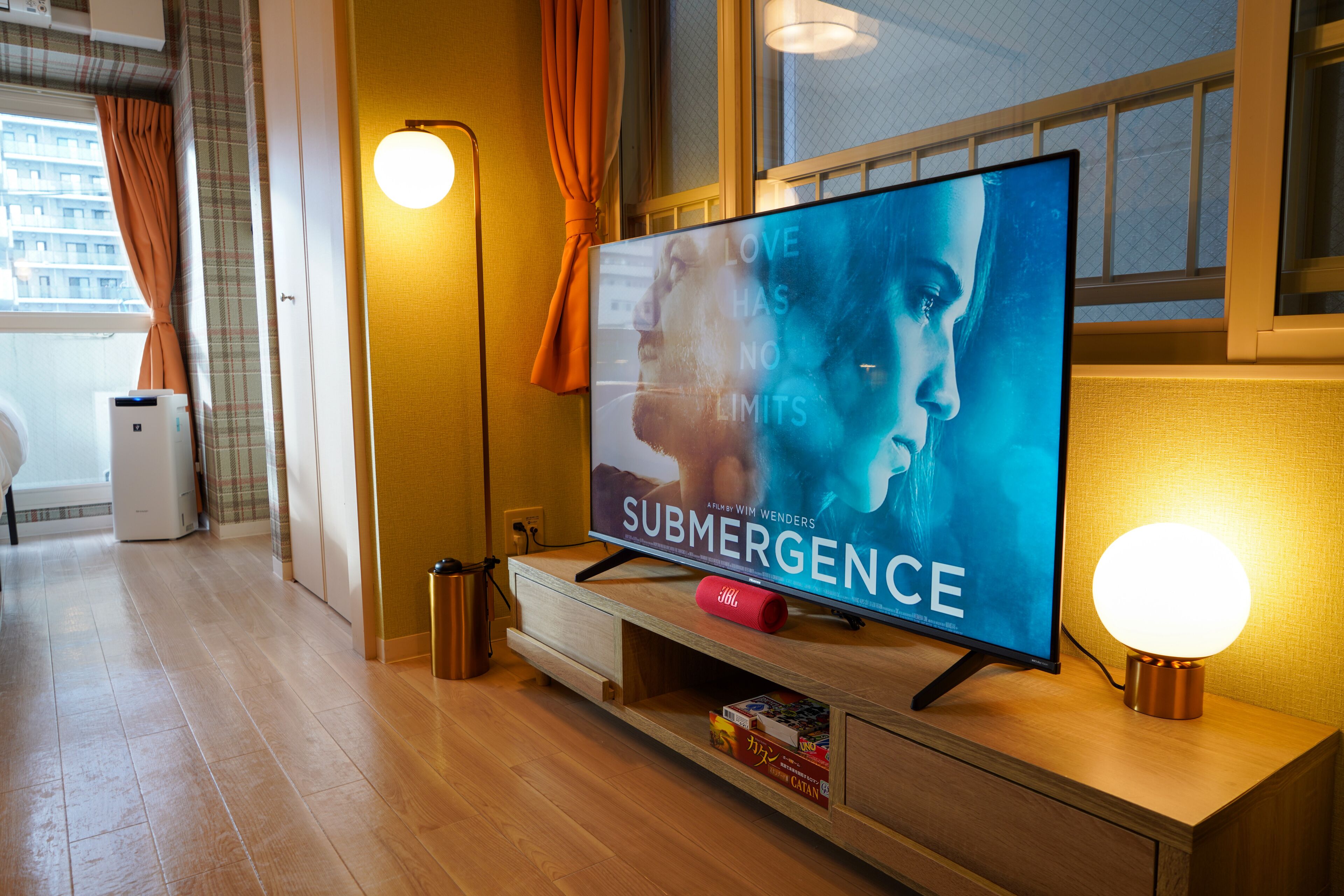 Una habitación de hotel moderna con una televisión de pantalla plana sobre un mueble de madera, una pequeña mesa con una vela roja y una ventana con cortinas.