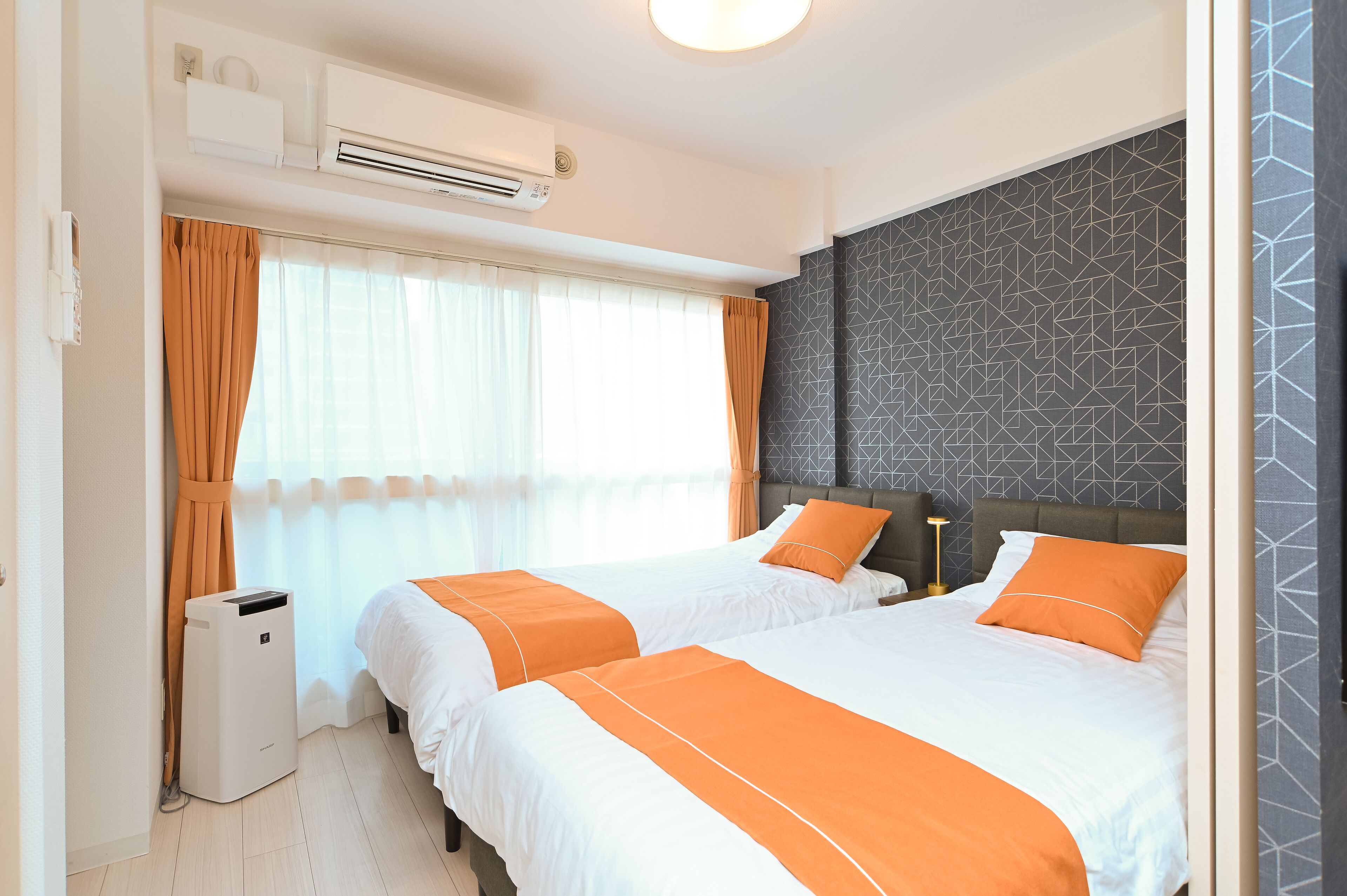 Une chambre d’hôtel avec deux lits, un appareil de climatisation, une poubelle et une fenêtre avec des rideaux.