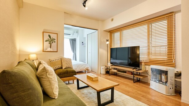 A modern living room with a green sofa, a wooden coffee table, a flat-screen TV, and a large window with blinds.