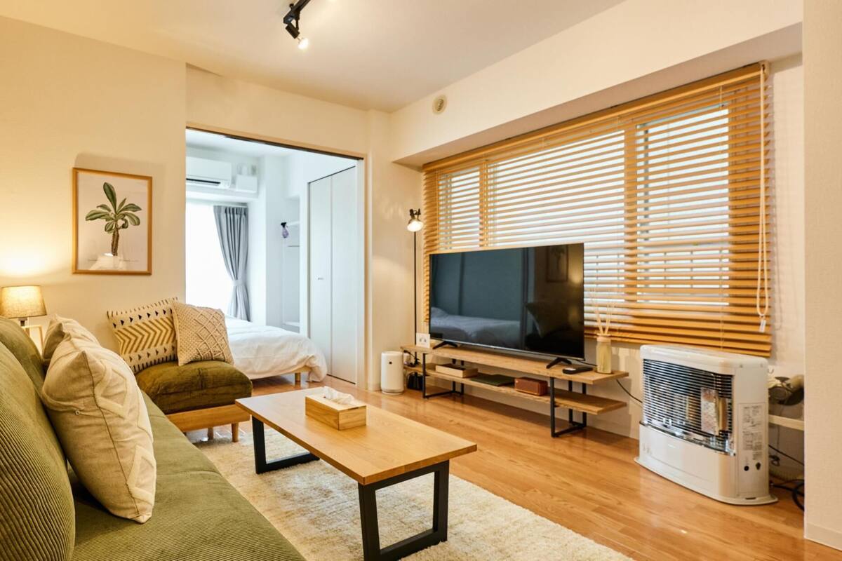 A modern living room with a green sofa, a wooden coffee table, a flat-screen TV, and a large window with blinds.