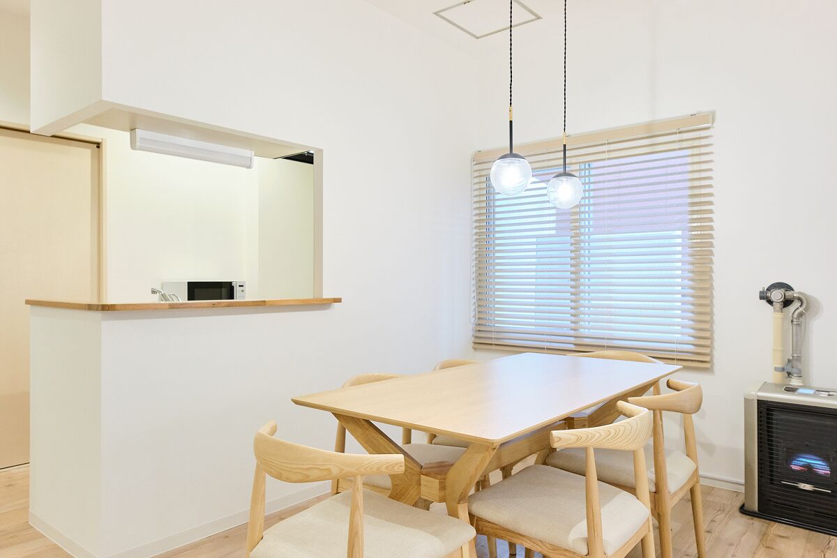 A dining area with a wooden table and chairs, a hanging light fixture, a window with blinds, and a built-in heater.