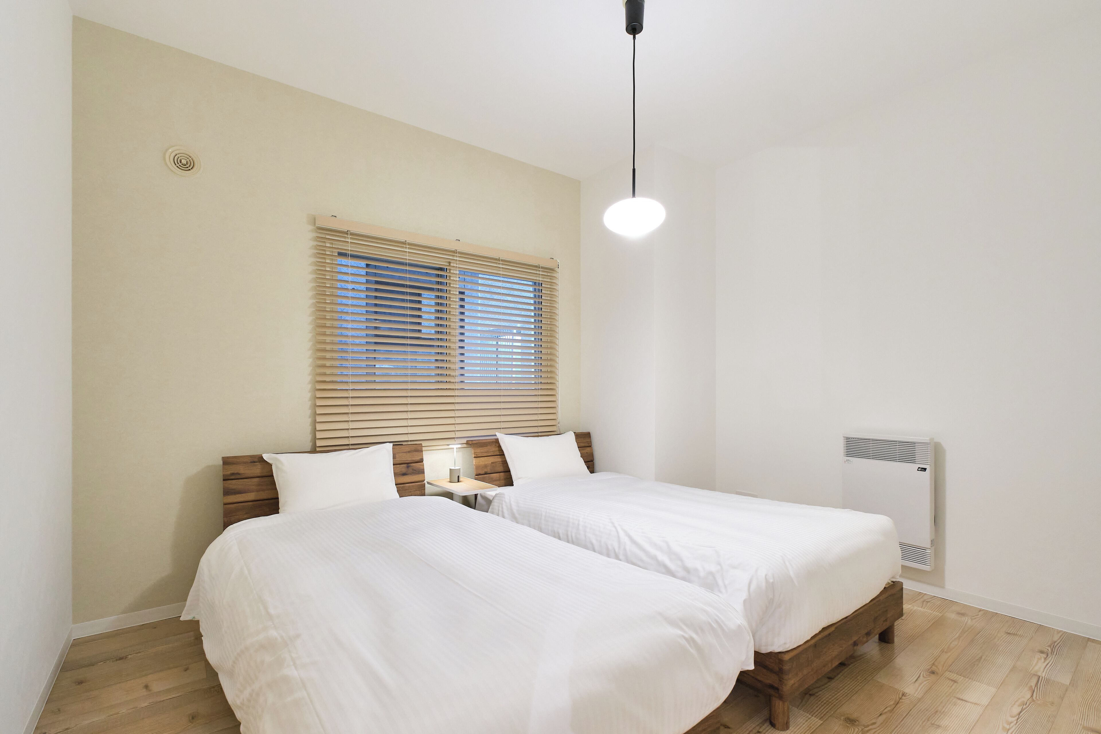 A bedroom with two beds, a window with blinds, a bedside table with a lamp, and a wall-mounted heater.