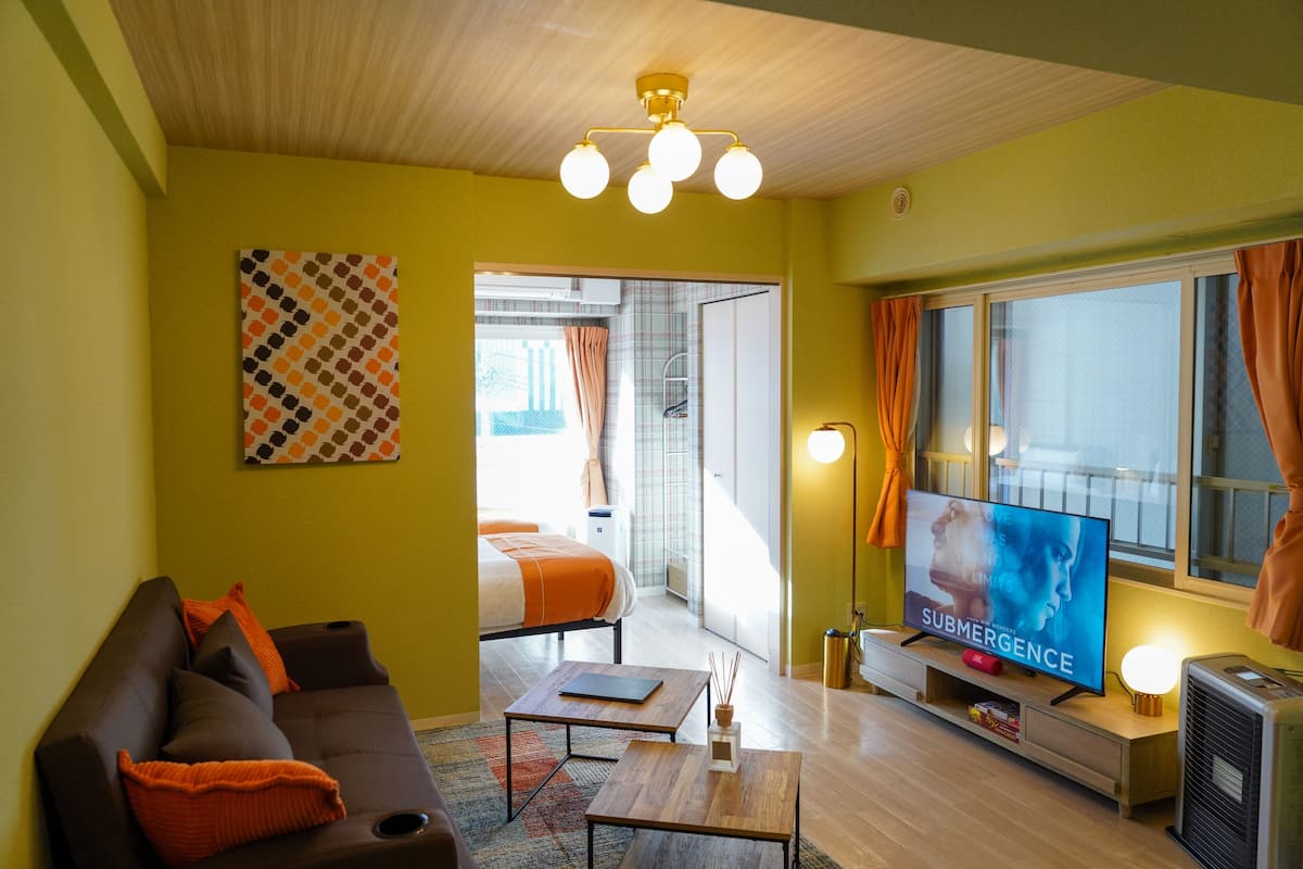 A modern living room with a sofa, coffee table, and TV. There is a bedroom visible through an open door.