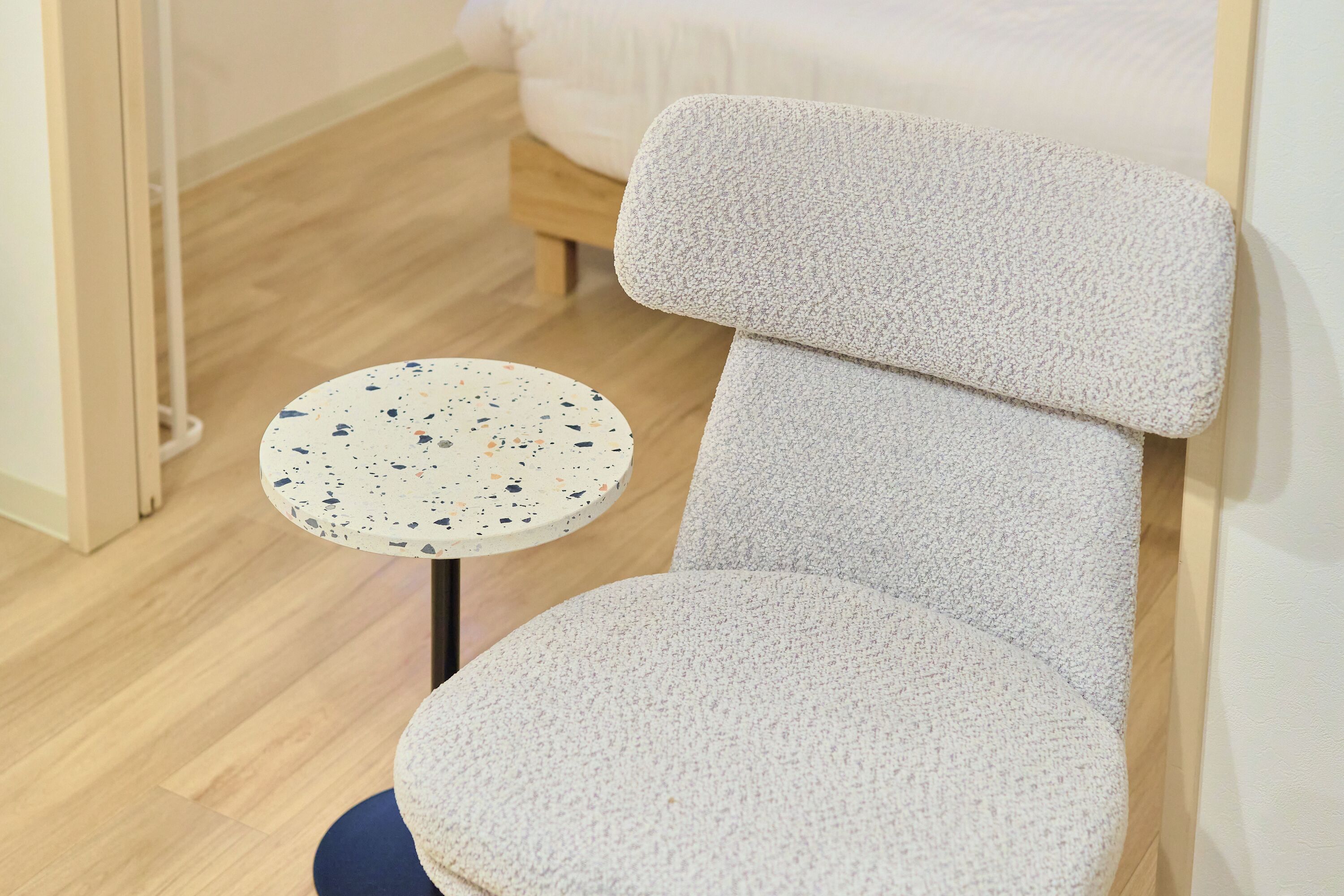 A room with a bed, a round table with a speckled tabletop, and a chair with a textured backrest.