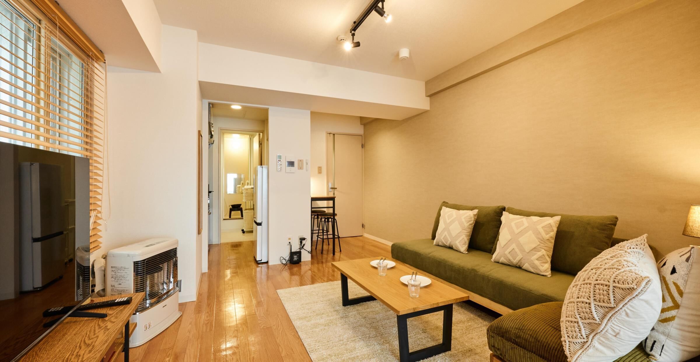 A modern living room with a sofa, coffee table, and a TV. There is a kitchen area in the background with a refrigerator and a microwave.