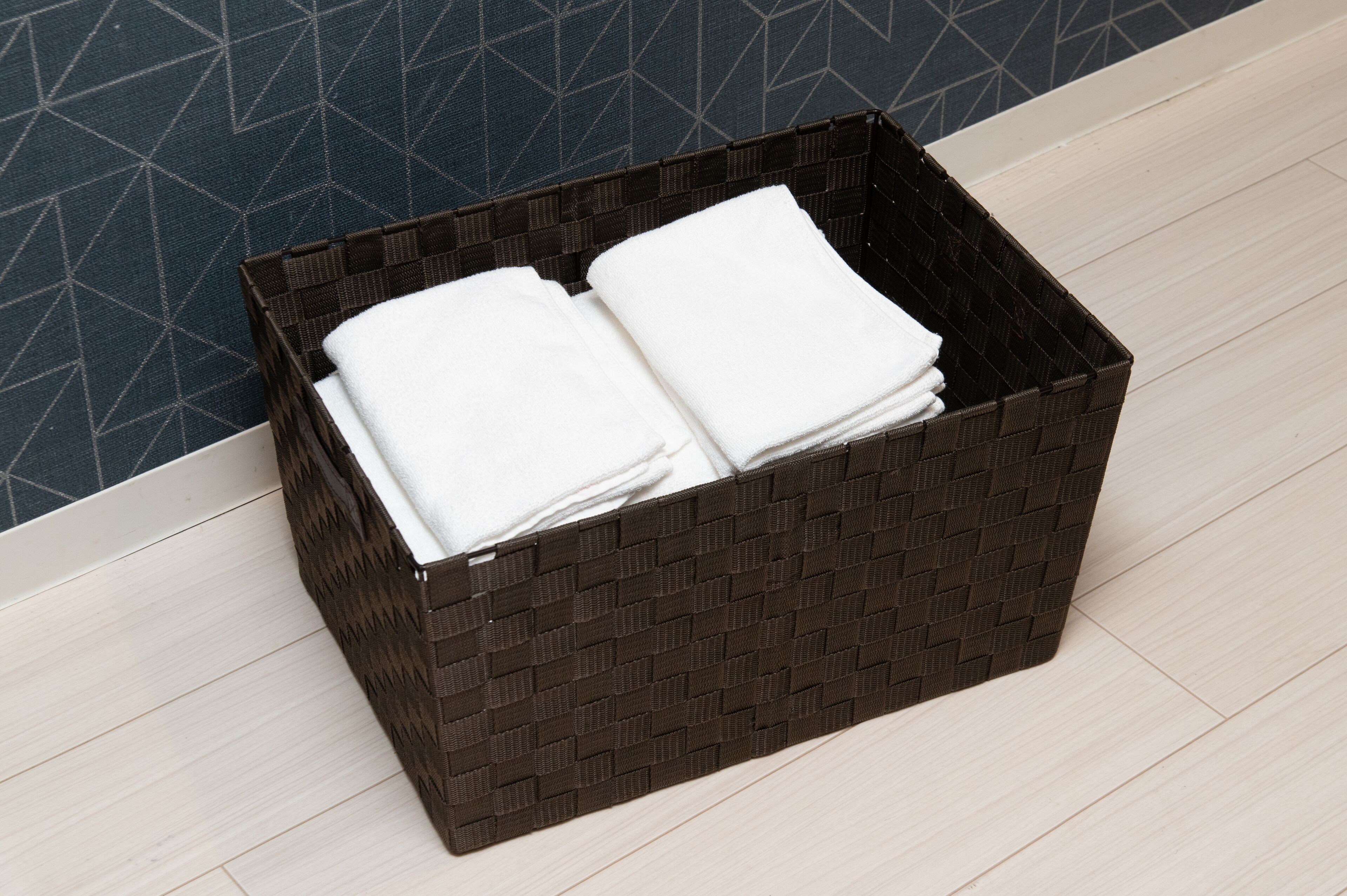 A dark woven basket with folded white towels on a light wooden floor next to a blue patterned wall.