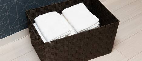 A dark woven basket with folded white towels on a light wooden floor next to a blue patterned wall.