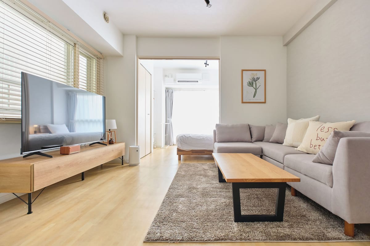 A modern living room with a sofa, a coffee table, a TV on a wooden stand, and a large window with blinds.