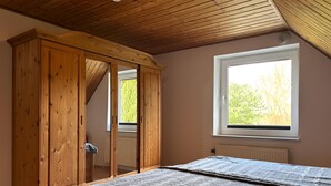 A bedroom with a wooden wardrobe, a bed with a grey blanket, a window with a view of trees, and a wooden ceiling. - Ferienwohnung Teschow (Teterow)